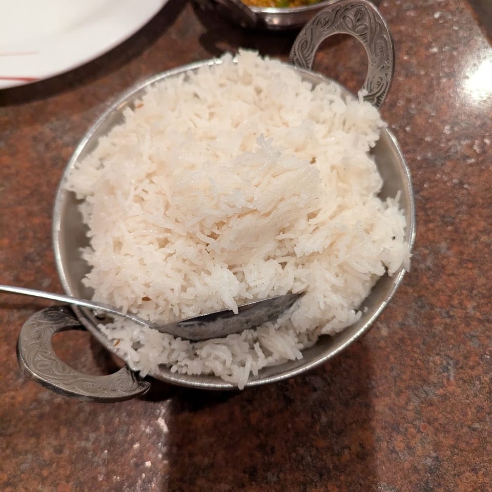 White rice in a decorative metal bowl with a spoon, with curry in the background.